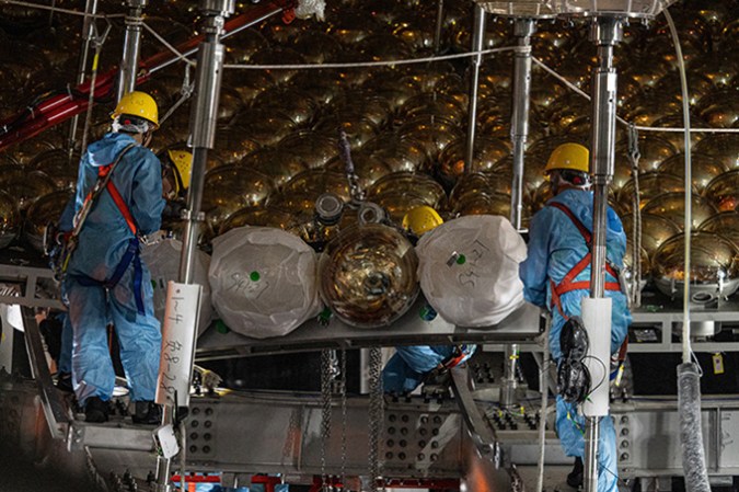 121624_ec_neutrino-detector_slide3-1 Four workers with hard hats and overalls are surrounded by golden orbs.